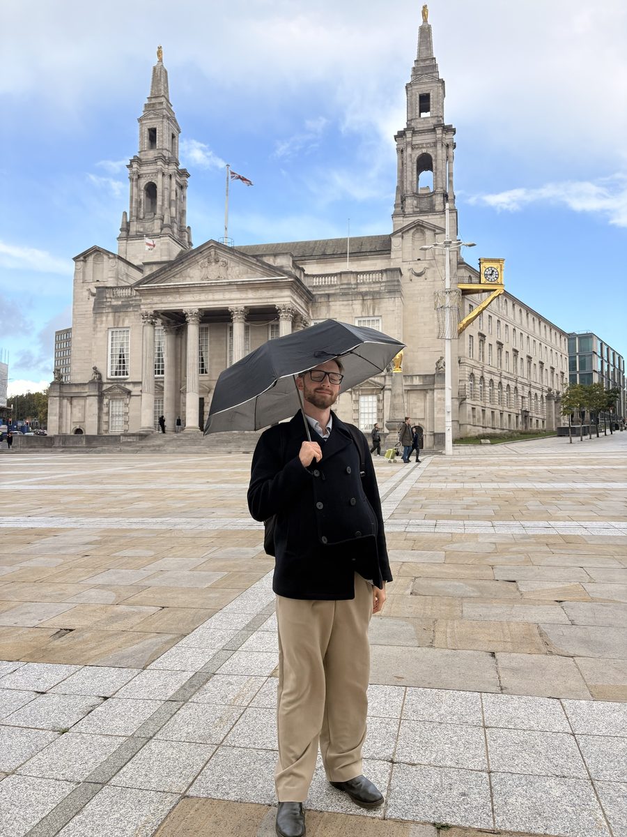 Mateusz Tomczak standing under an umbrella in a navy coat on a city square